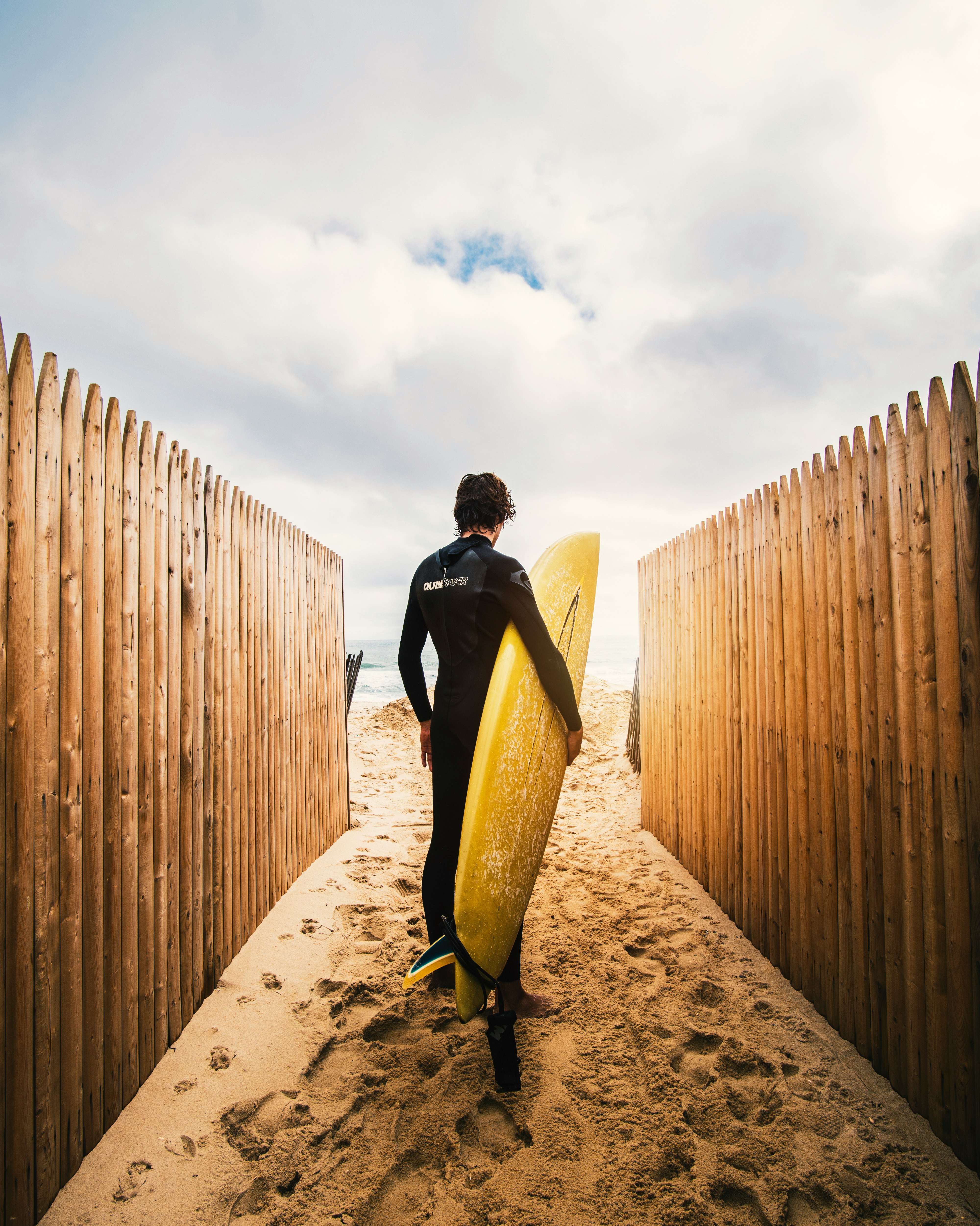 person standing between privacy fences while holding yellow surfboard during daytime